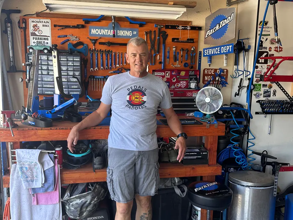 The owner standing at his garage workbench, with the full pegboard of bicycle tools, a Park Tool repair stand and a shelf of parts drawers behind him.