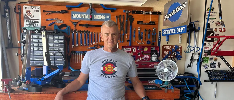 The garage workshop: a pegboard wall of Park Tool wrenches, hex keys and cable cutters above a wooden bench, parts drawers and a repair stand to the left, and the owner standing at the bench with the garage door open behind him.