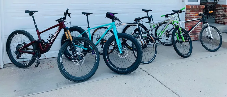 Six bikes lined up along the garage door after work: a maroon full-suspension mountain bike, a turquoise hardtail, a fat bike, a black road bike and two more hardtails.