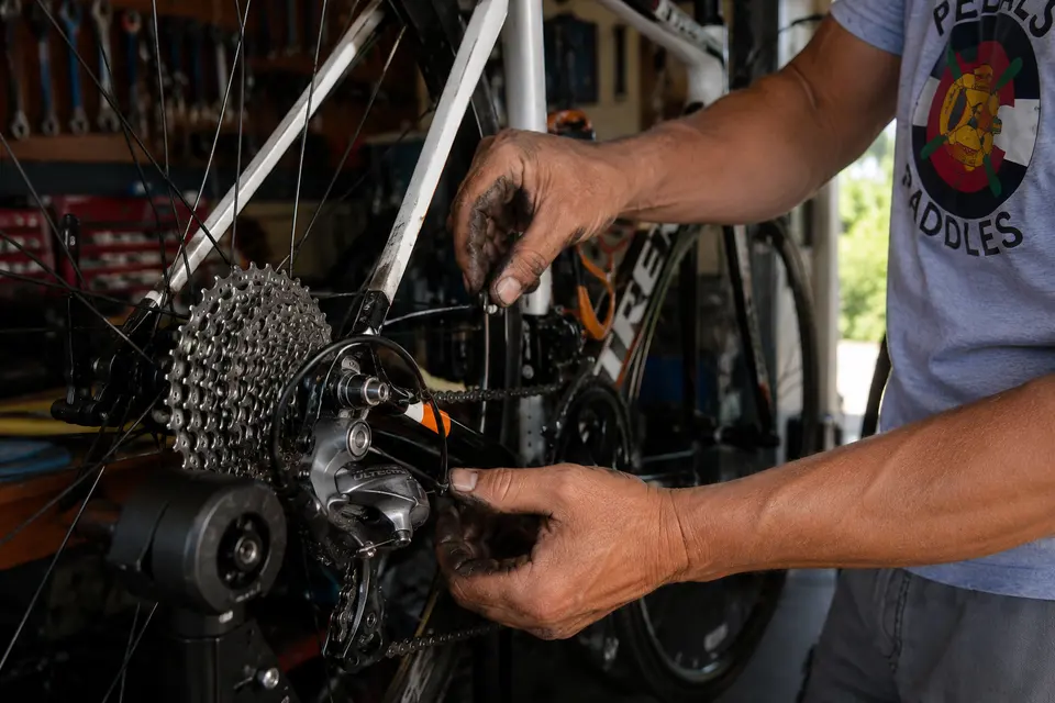 Two greasy hands on the rear derailleur and cassette of a bike in the stand, chain and spokes in sharp focus, the tool board out of focus behind.