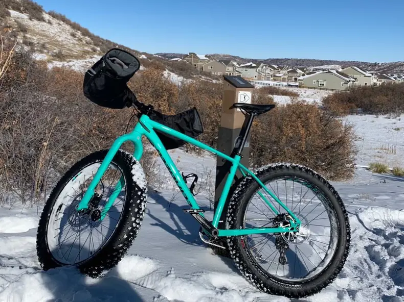 A turquoise fat bike with oversized studded tyres parked in packed snow on a Colorado trail, with a frosted hillside and houses in the distance.
