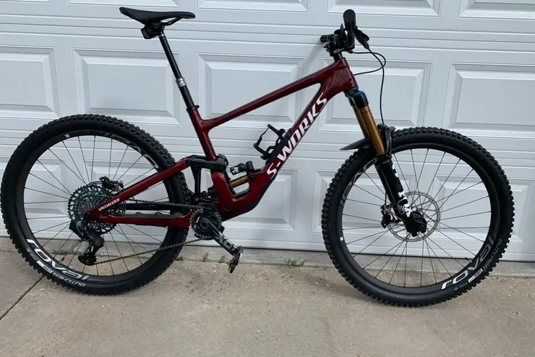 A deep red S-Works full-suspension mountain bike standing on a driveway against a white garage door, with knobby tyres and carbon Roval wheels.