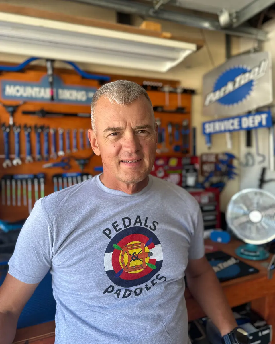 The owner, chest-up in his garage workshop, wearing a grey Pedals & Paddles t-shirt, with the tool wall and a Park Tool sign out of focus behind him.
