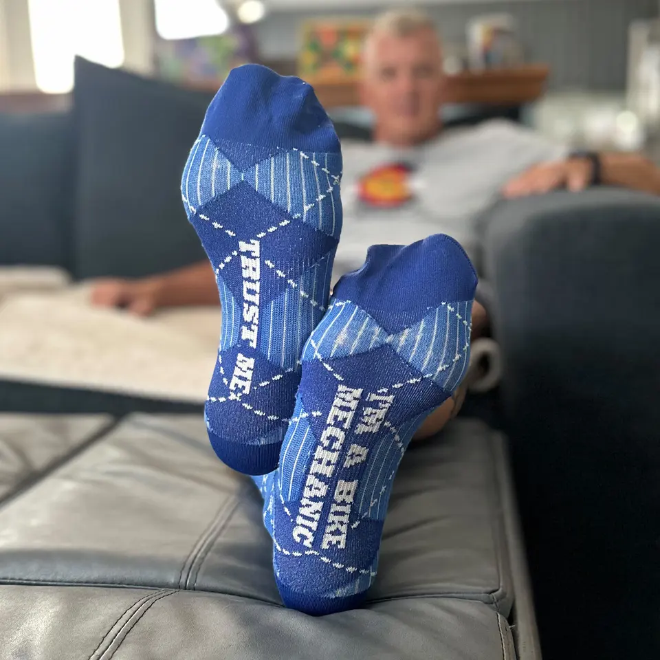 A pair of blue argyle socks with “Trust me, I’m a bike mechanic” knitted across the soles, feet up on an ottoman.